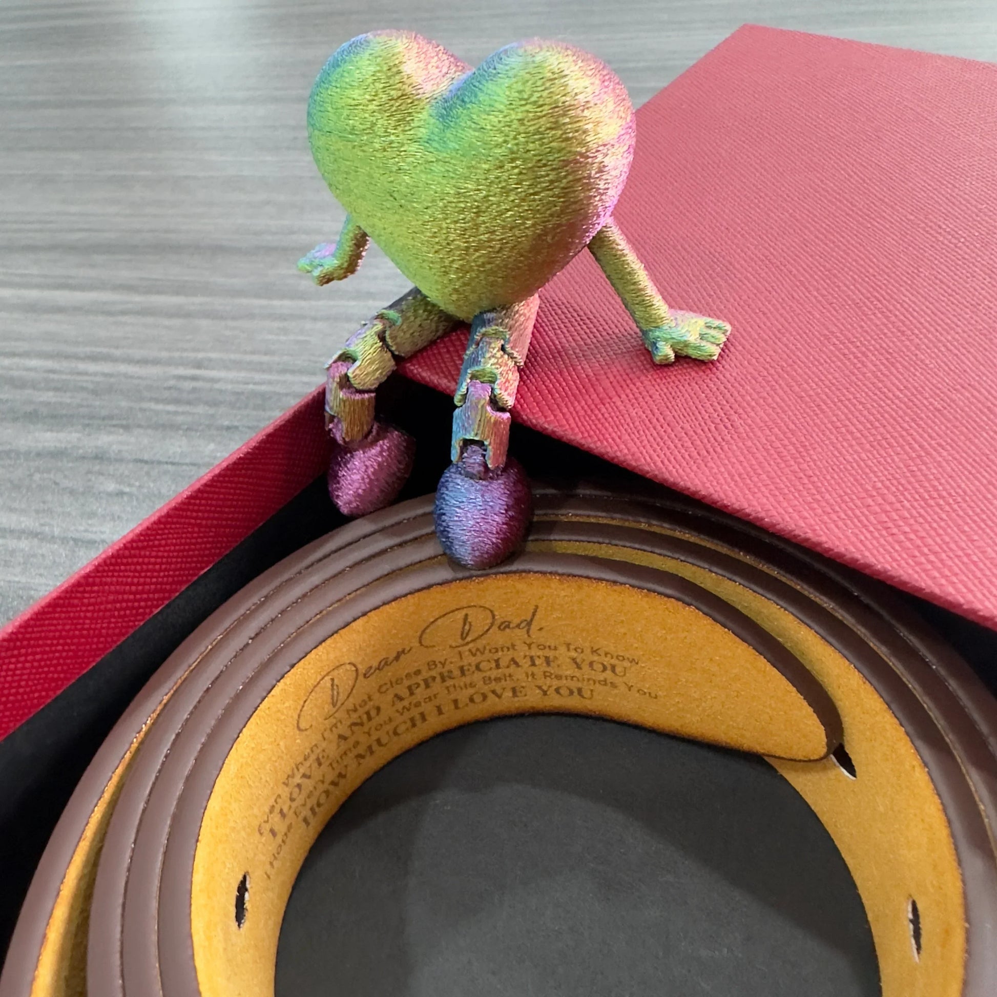 Brown leather belt with engraved message, colorful heart figurine, and red gift box on table.