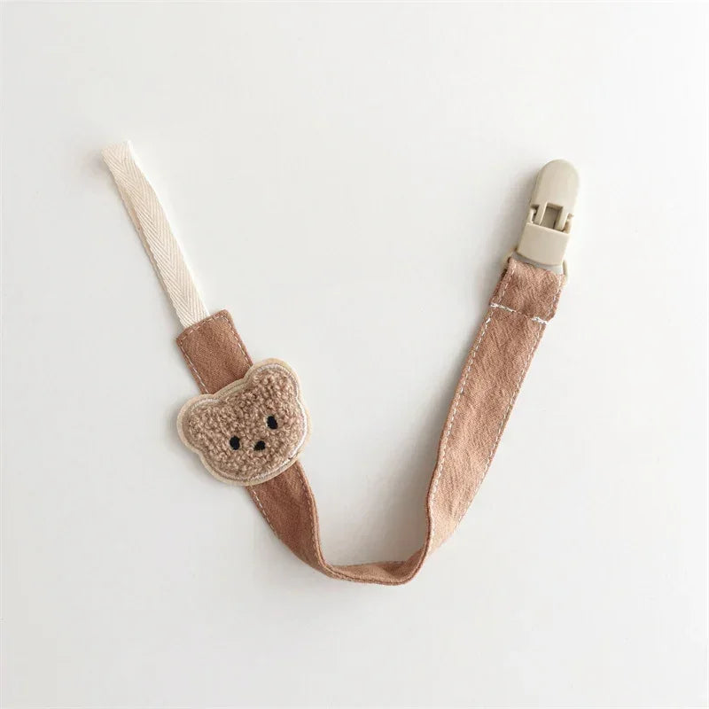 Brown fabric pacifier clip with soft bear patch and beige plastic clasp, minimalist design