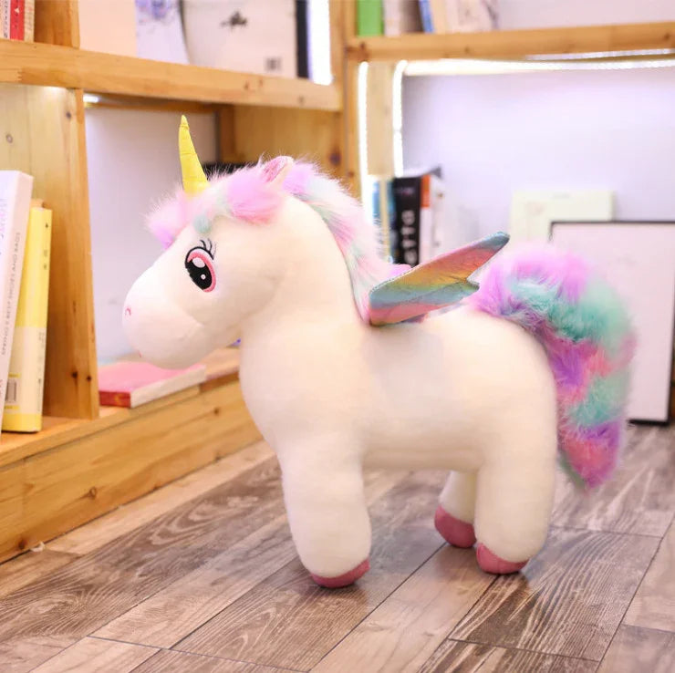 Plush white unicorn toy with rainbow mane, tail, wings, and yellow horn on wooden floor
