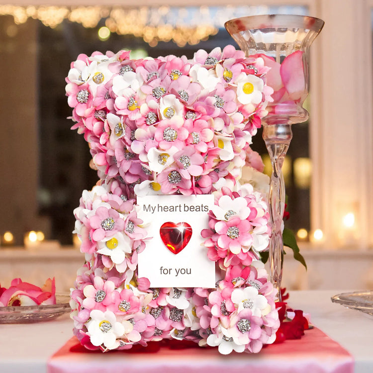 Decorative pink and white artificial flower bear with sparkling accents, heart-shaped note on display.
