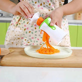 Person wearing a colorful apron with leaf print, using a white and green spiralizer kitchen tool to make carrot noodles, apron fits loose and casual.