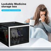 Woman in grey knit sweater with blanket, holding a mug and tissue, beside a lockable medicine storage box on a table.