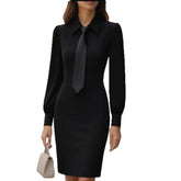 Woman in fitted black long sleeve shirt dress with tie, holding a beige handbag