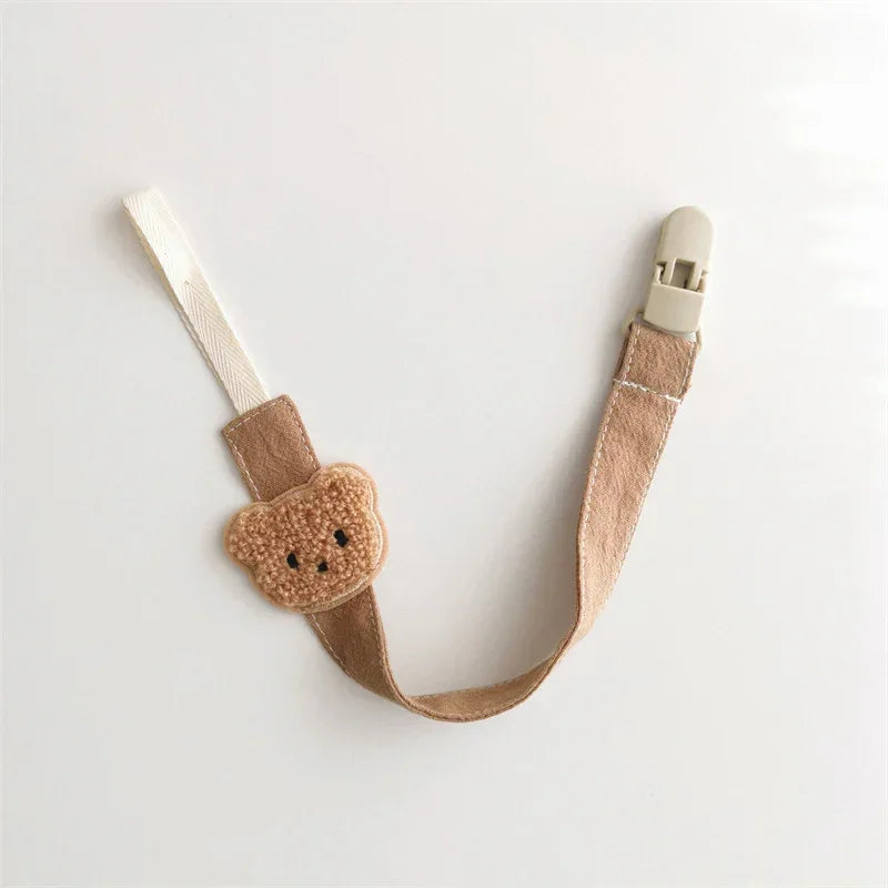 Beige fabric pacifier clip with plush bear face, metal clasp, and soft cotton strap