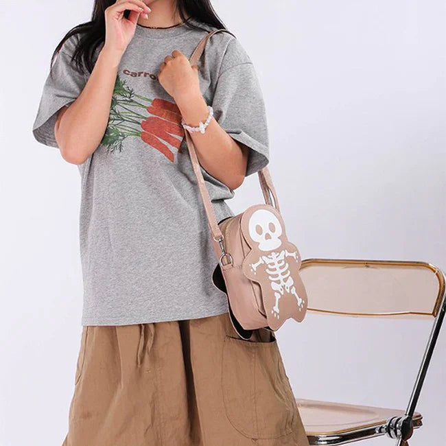 Woman in oversized grey carrot graphic t-shirt and tan skirt, holding tan skeleton crossbody bag