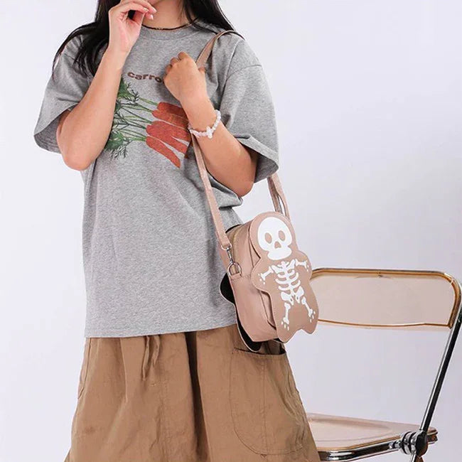 Woman in oversized grey carrot graphic t-shirt and tan skirt, holding tan skeleton crossbody bag