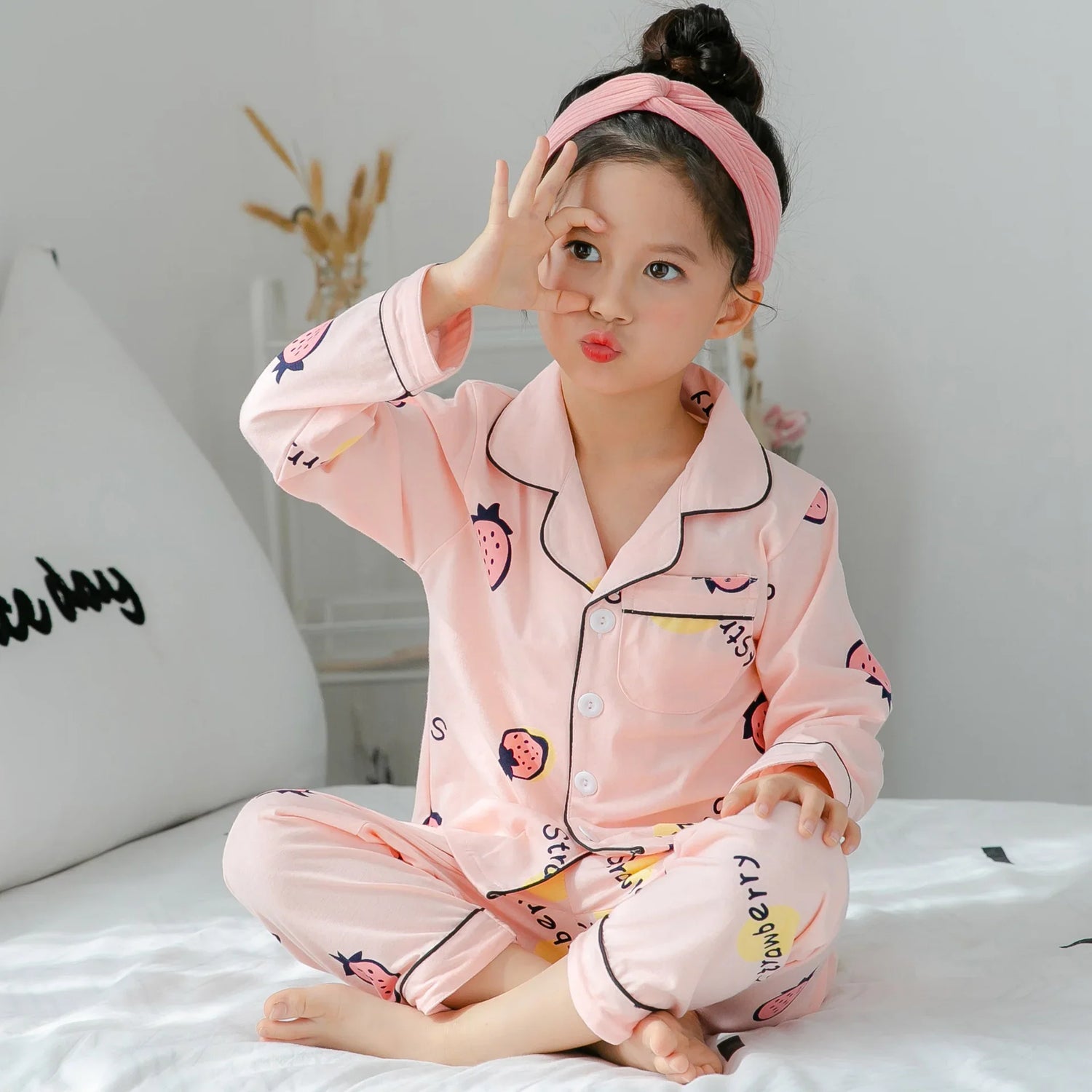 Child in light pink cartoon strawberry print pajamas with black trim, relaxed fit, sitting on bed