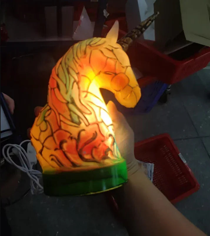 Hand holding a glowing unicorn head lamp with multicolor stained glass look and green base.