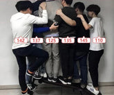 Group of young adults wearing casual streetwear, layered jackets, hoodies, and sneakers in neutral and dark colors, all standing together closely on a skateboard indoors.