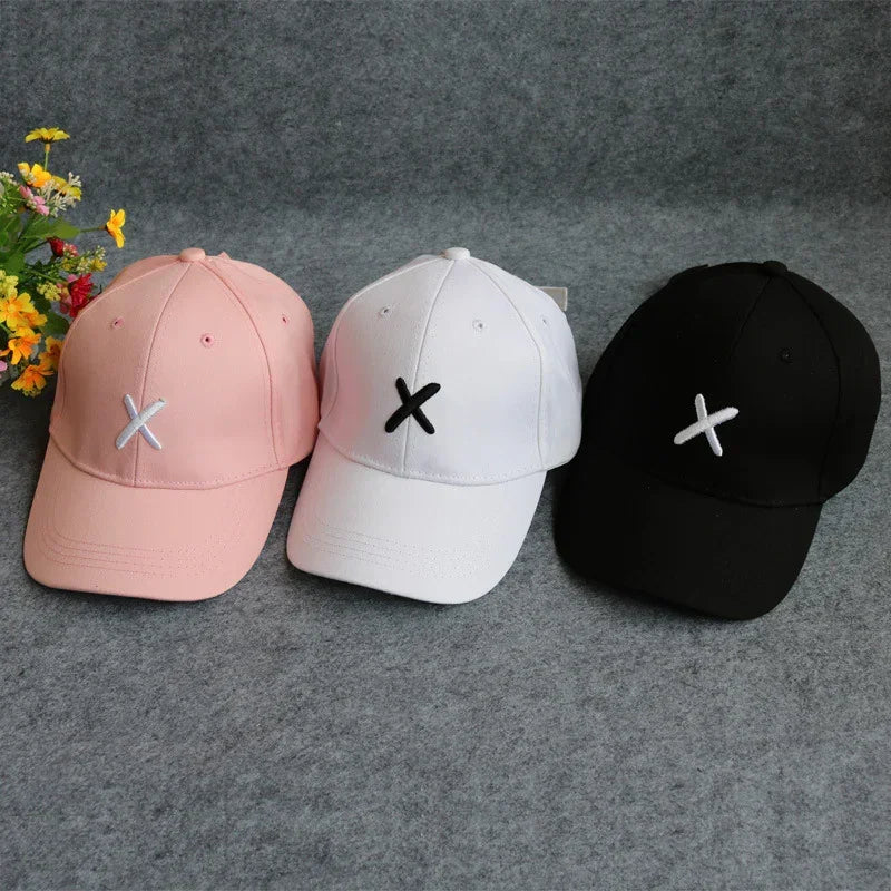 Three cotton baseball caps in pink, white, and black with embroidered X design, minimalist streetwear style.