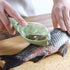 Plastic fish scaler tool removing scales from raw fish on wooden cutting board