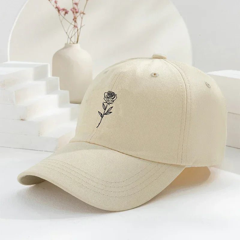 Beige cotton baseball cap with black embroidered rose graphic, curved brim, relaxed fit.