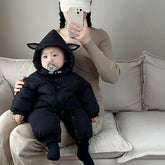 Baby in black hooded puffer jumpsuit with animal ears, sitting on a beige sofa