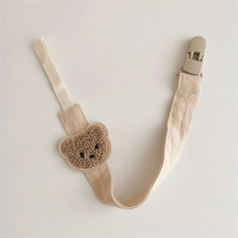 Beige fabric pacifier clip with cute teddy bear patch and plastic clasp, minimalist style.