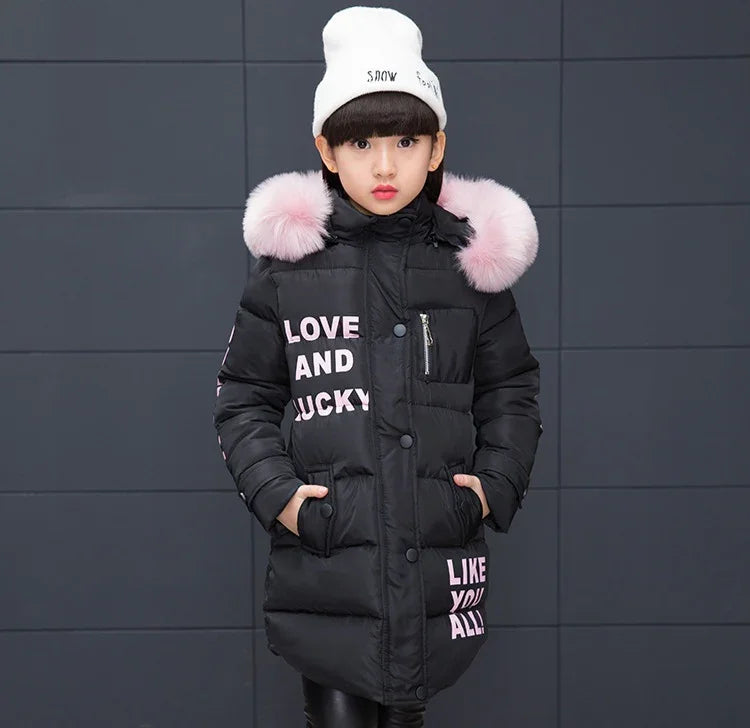 Girl wearing long black puffer jacket with pink faux fur hood, white beanie, and letter print.
