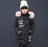 Girl wearing long black puffer jacket with pink faux fur hood, white beanie, and letter print.