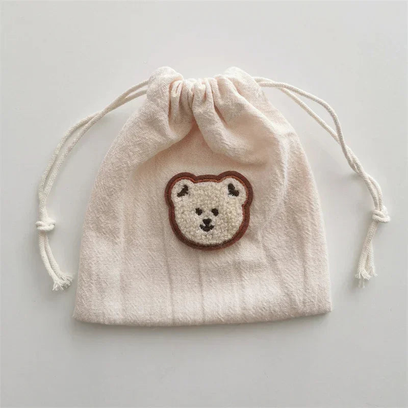 Beige cotton drawstring pouch with embroidered bear face patch, soft textured fabric.