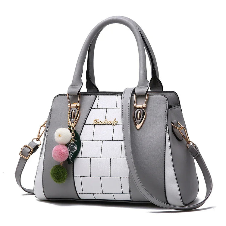 Gray and white structured handbag with geometric panels, top handles, and colorful pompom charm.