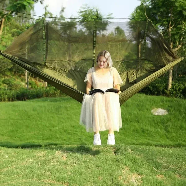 Woman in a sheer beige tulle dress and white sneakers reading on a green hammock outdoors