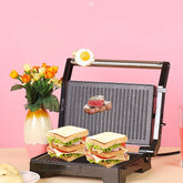 Double-decker grilled sandwiches with lettuce and deli meat on a black panini press, pink background.