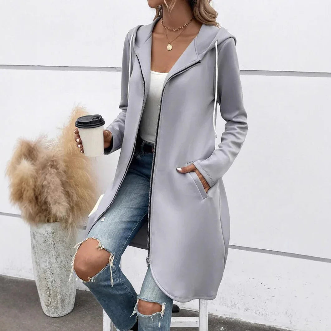 Woman wearing light gray hooded zip-up longline jacket, white tank top, and ripped blue jeans.
