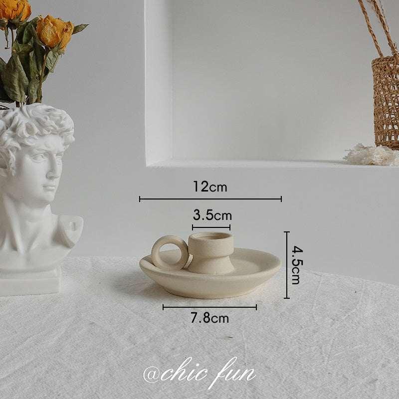Minimalist beige ceramic candle holder with ring handle, placed on a white table, shown with dimensions 12cm wide, 4.5cm tall.
