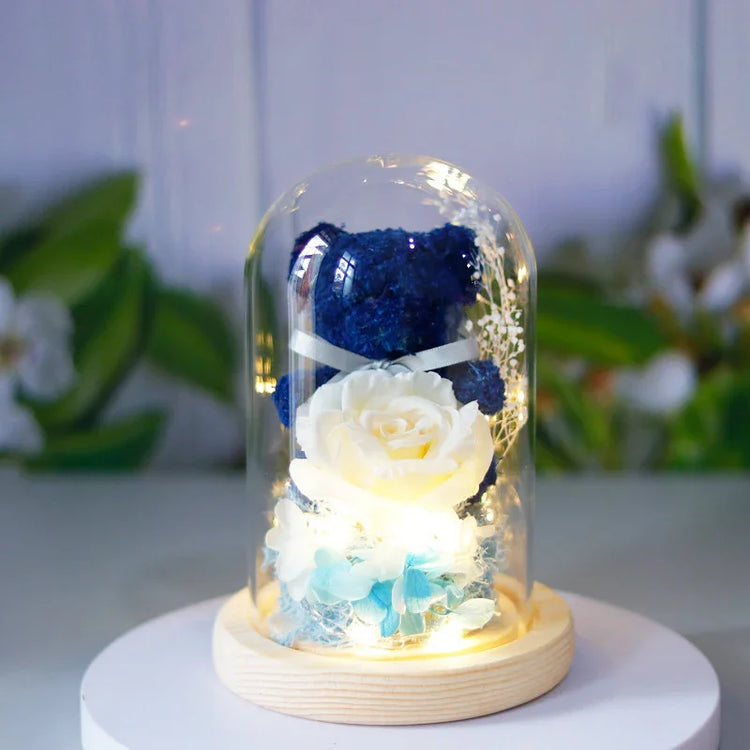 Blue plush bear with white rose and blue flowers in a glass dome with LED lights