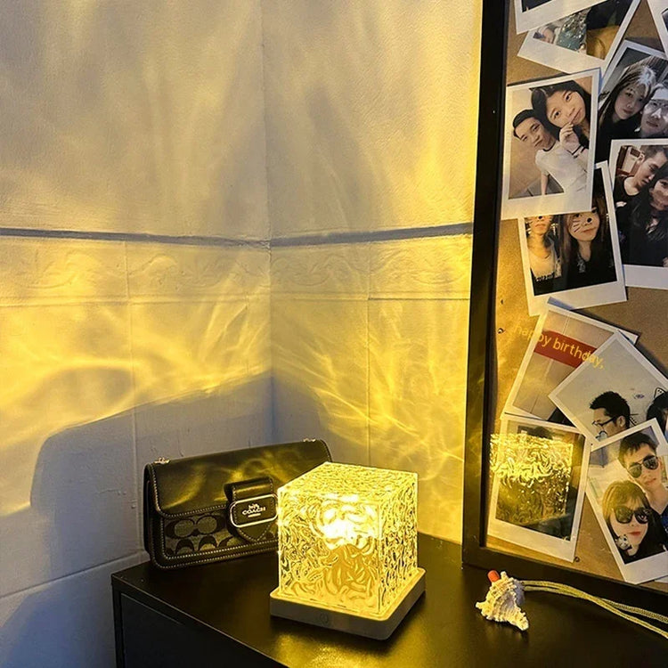 Cube-shaped textured glass lamp with warm yellow light on a black table near a photo wall.