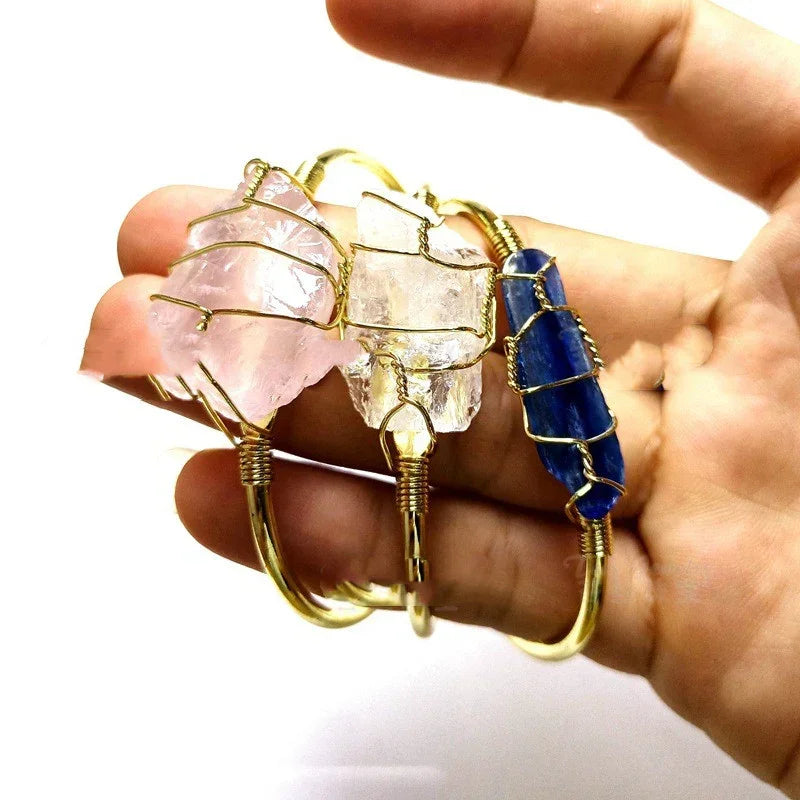 Hand holding wire-wrapped crystal bracelets in pink, clear, and blue hues, gold metal accents