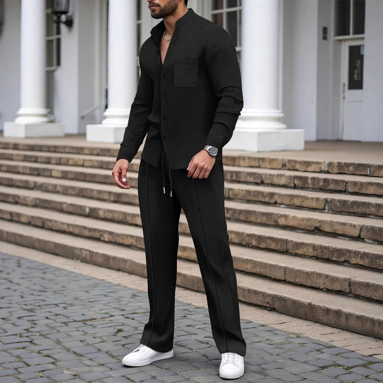 Man wearing black textured button-up shirt and matching pants in a relaxed fit, styled with white sneakers.