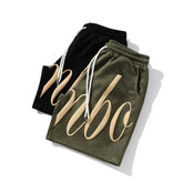 Two pairs of streetwear shorts, olive green and black, bold graphic lettering, elastic waist, drawstring, relaxed fit.