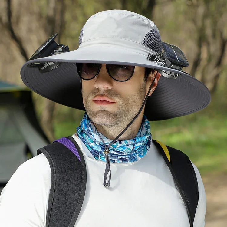 Man wearing a wide-brimmed sun hat with solar-powered fans, white shirt, patterned neck gaiter, and backpack outdoors.