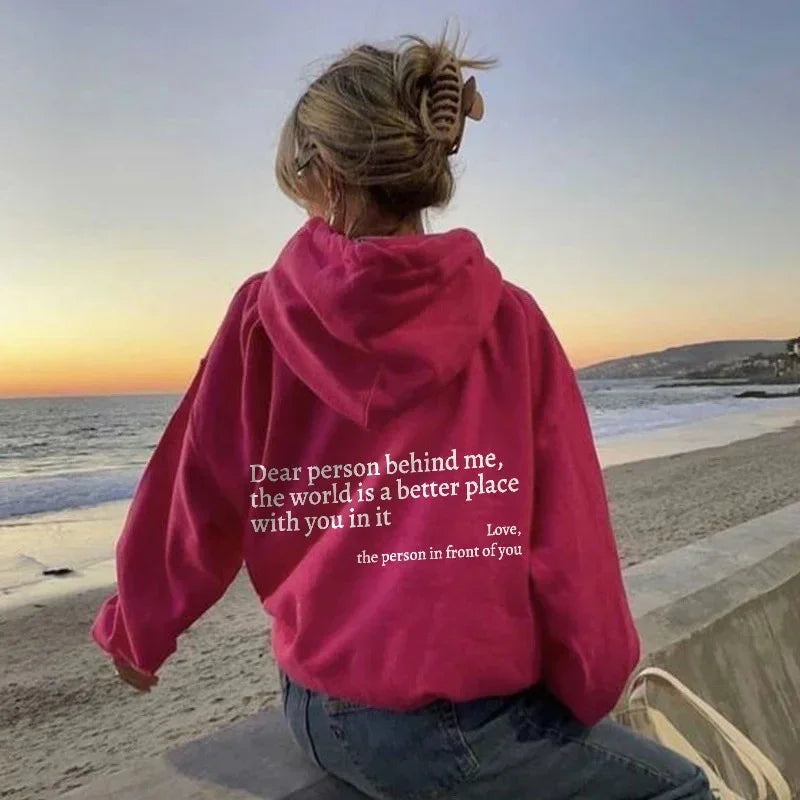Woman in oversized pink hoodie with inspirational text, casual fit, sitting by the beach at sunset
