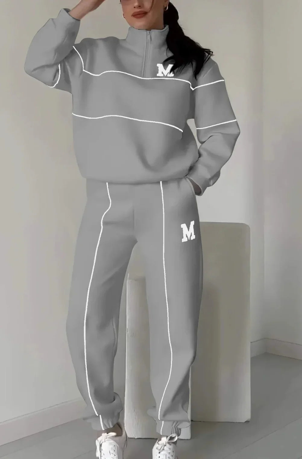 Woman wearing a gray oversized tracksuit set with white piping, relaxed fit, and letter M detail.