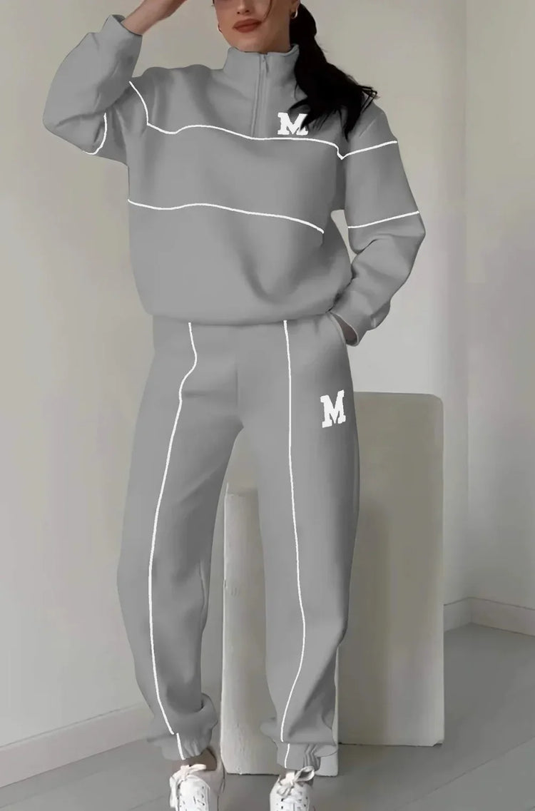 Woman wearing a gray oversized tracksuit set with white piping, relaxed fit, and letter M detail.