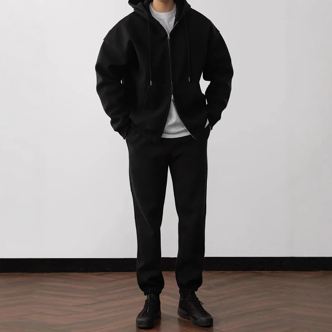Man wearing black oversized zip-up hoodie, matching jogger pants, and black sneakers, relaxed fit.