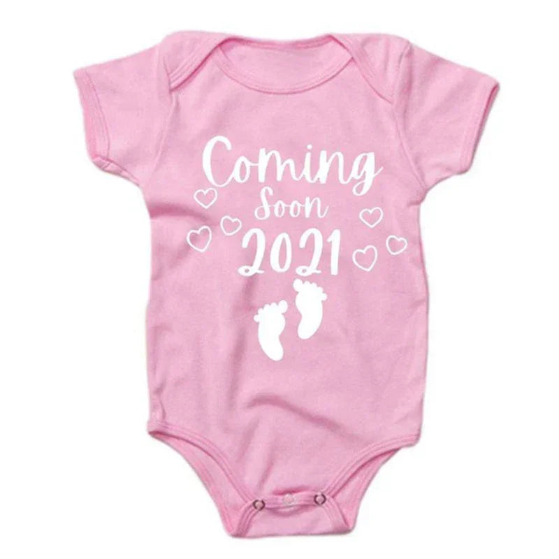 Pink short-sleeve cotton baby onesie with 'Coming Soon 2021' text and heart print, snap closure.