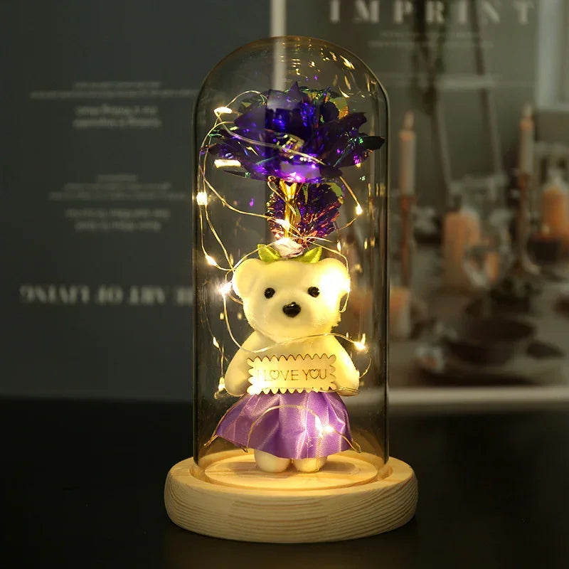 White teddy bear in purple skirt with 'I LOVE YOU' sign under glass dome, fairy lights, floral topper.