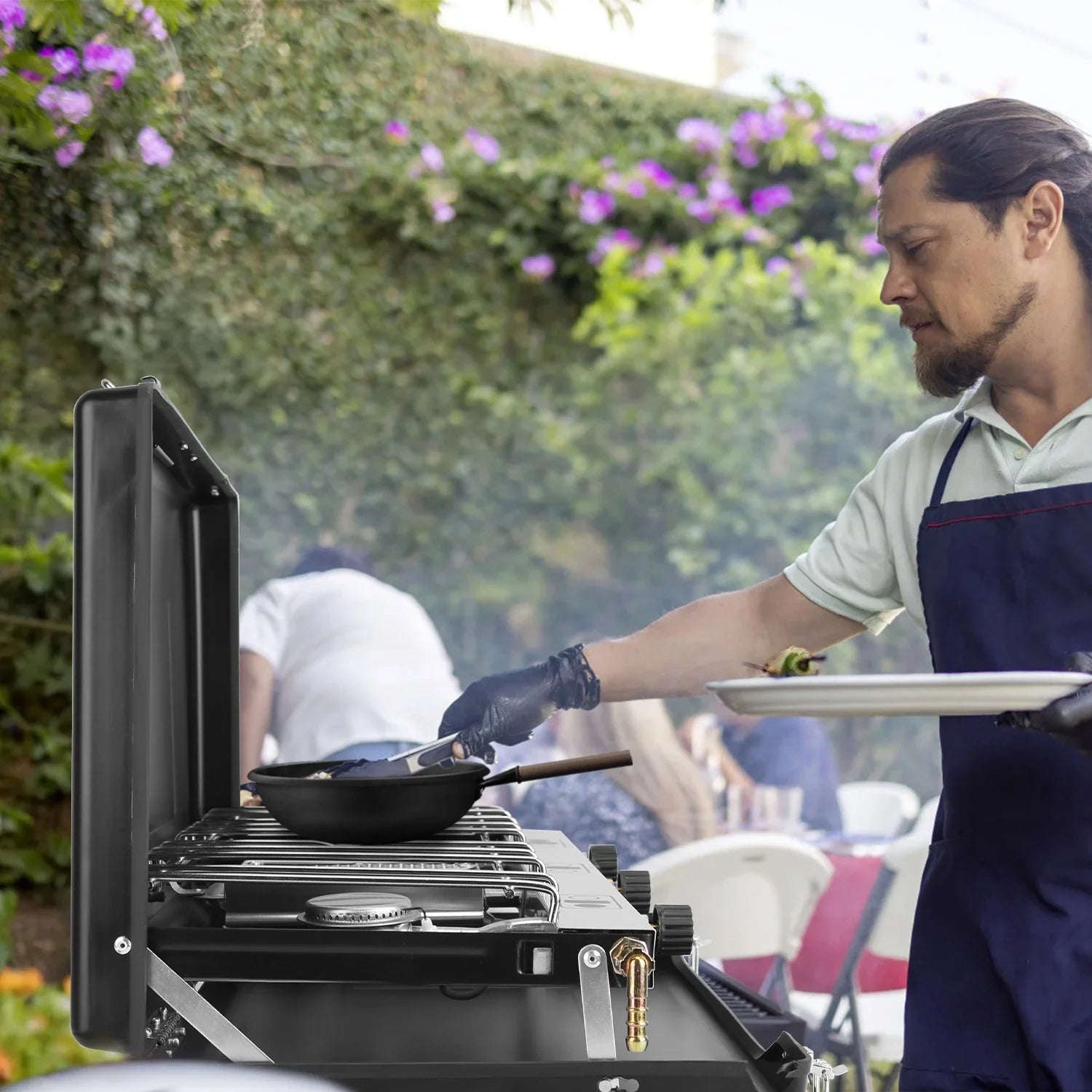 Man in a short sleeve polo and navy apron grilling outdoors, wearing black gloves