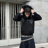 Man wearing a black zip-up hoodie with faux fur hood, gothic print, and baggy jeans with studded belts