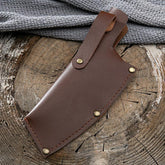 Brown leather sheath with brass rivets, laid on rustic wood next to a textured grey fabric
