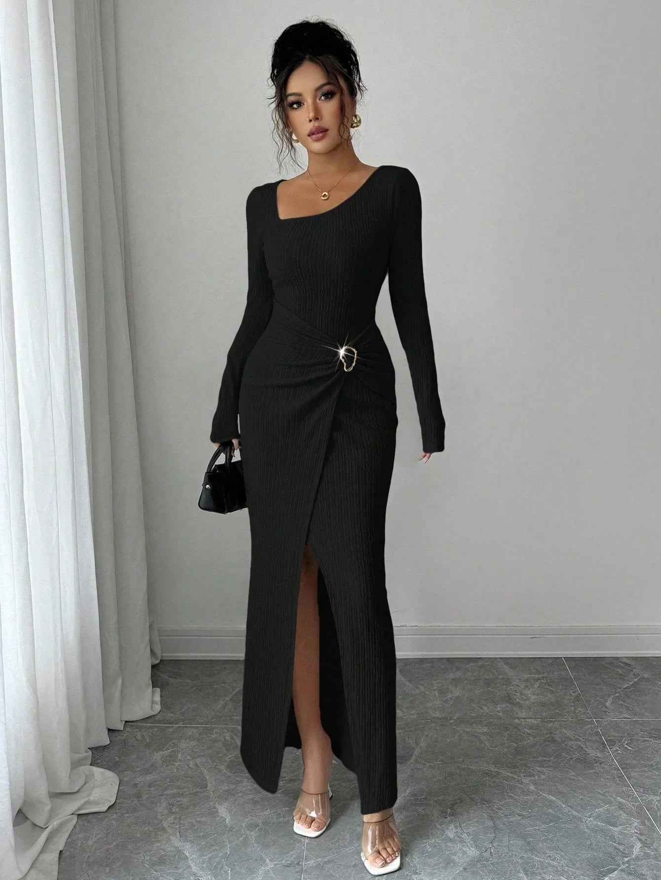 Woman wearing a long black ribbed bodycon dress with front slit, asymmetrical neckline, and metal ring detail, styled with clear heels and a small handbag.