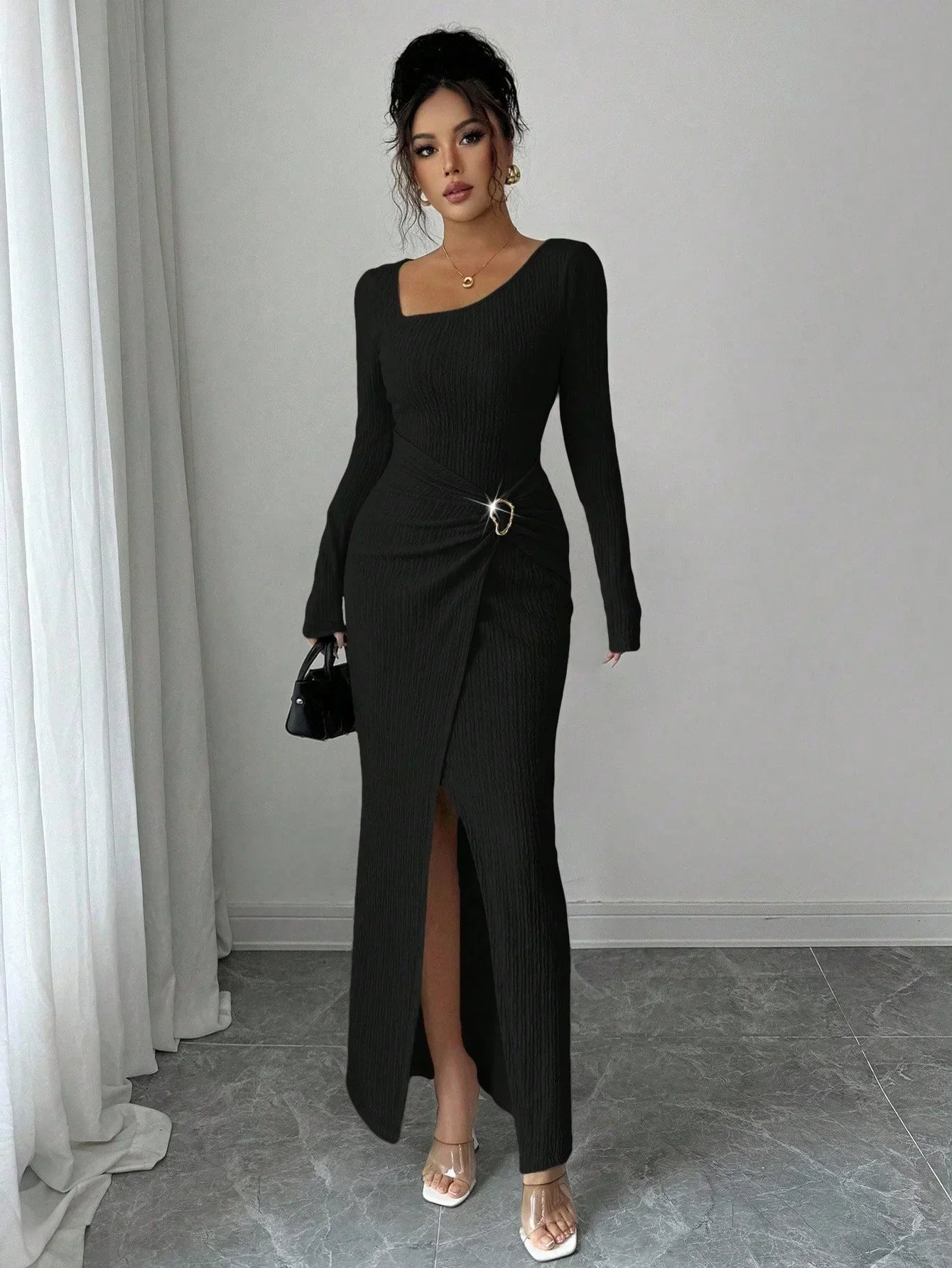 Woman wearing a long black ribbed bodycon dress with front slit, asymmetrical neckline, and metal ring detail, styled with clear heels and a small handbag.