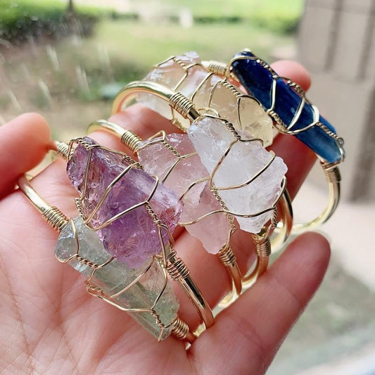 Hand holding gold wire-wrapped gemstone bracelets in assorted colors, chunky and modern style.