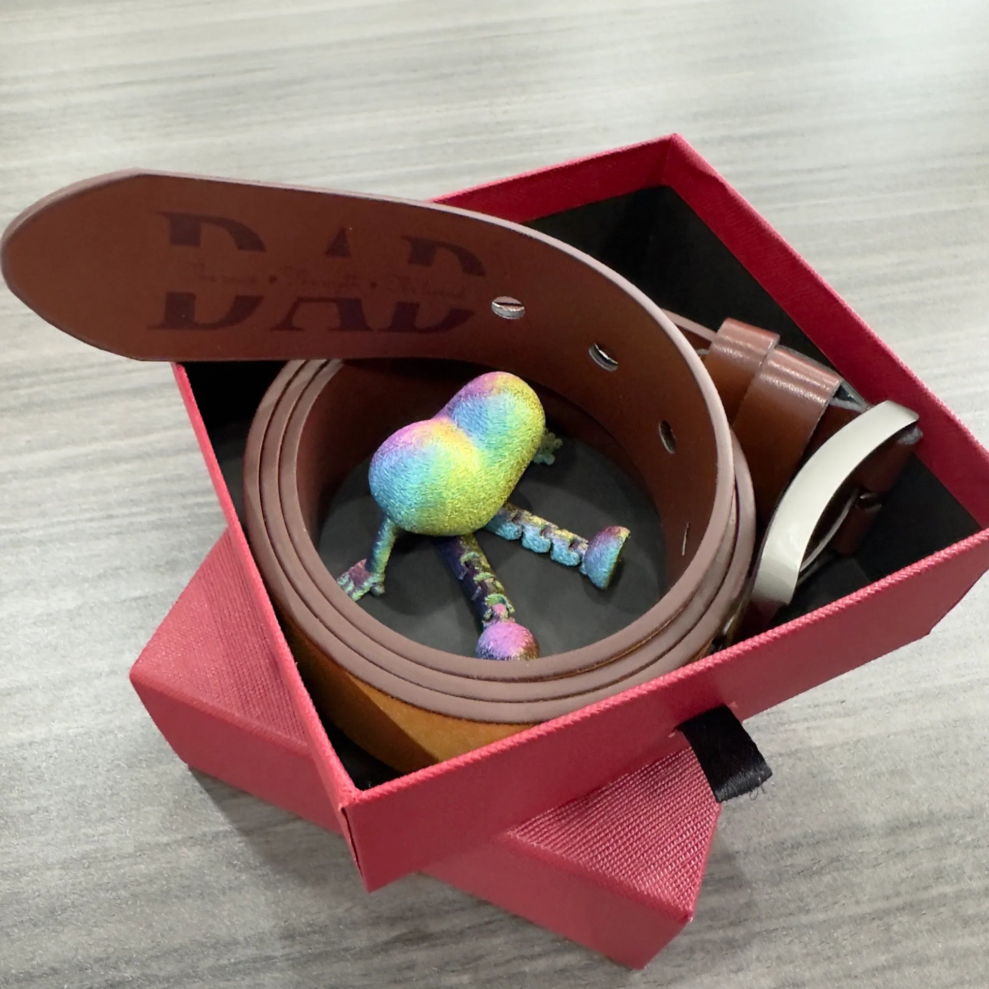 Brown leather belt with metallic buckle in a gift box, featuring a rainbow heart figurine.