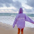 Person wearing oversized lavender hoodie with quote on back, standing on beach by ocean waves