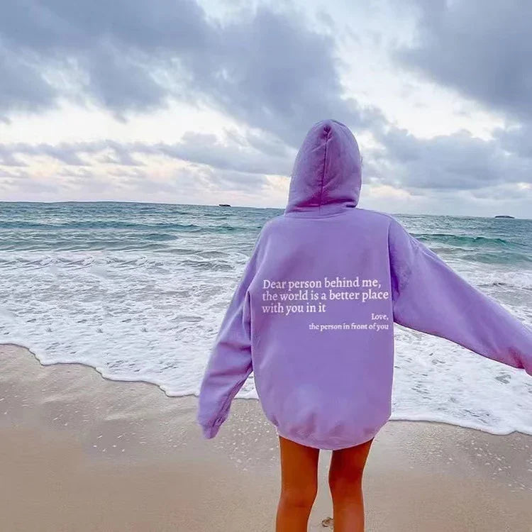 Person wearing oversized lavender hoodie with quote on back, standing on beach by ocean waves