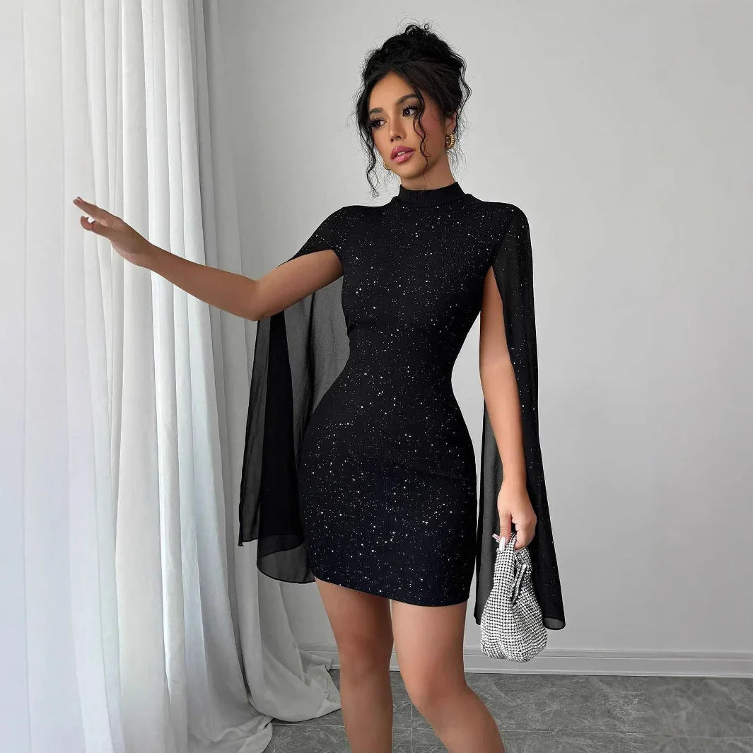 Woman wearing a fitted black glitter mini dress with sheer cape sleeves, holding a small purse