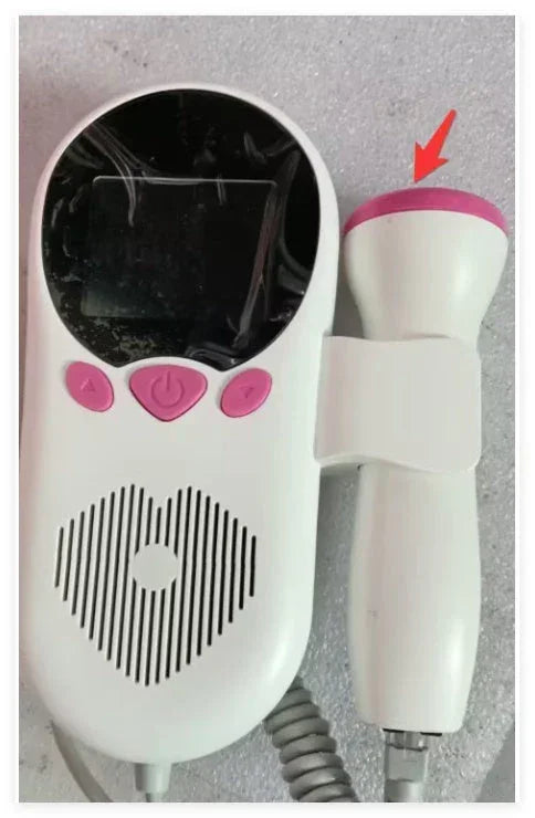 White handheld electronic device with pink buttons and accents, heart-shaped speaker grill, LCD screen, and detachable probe, shown on a textured grey surface.
