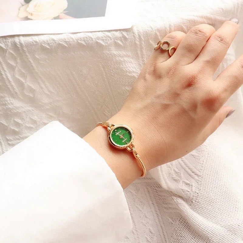 Gold bracelet watch with green face on wrist, paired with gold ring, white sleeve visible
